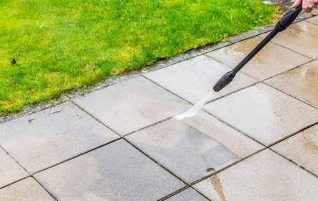 A person is using a pressure washer to clean outdoor patio tiles next to a patch of green grass, demonstrating techniques similar to commercial soft washing for gentle yet effective results.