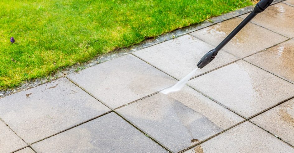 A person is using a pressure washer to clean outdoor patio tiles next to a patch of green grass, demonstrating techniques similar to commercial soft washing for gentle yet effective results.