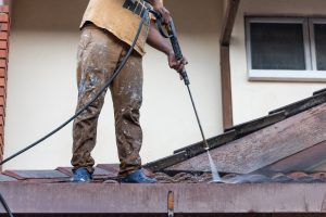 A person standing on a roof uses Residential Soft Washing techniques with a pressure washer to clean the tiles, as water spray is visible.