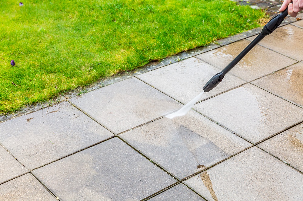 A person is using a pressure washer to clean outdoor patio tiles next to a patch of green grass, demonstrating techniques similar to commercial soft w